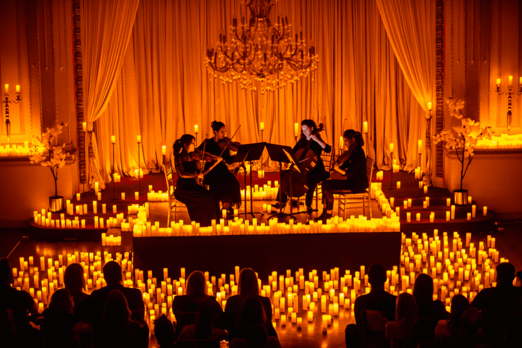 A string quartet performing on a stage covered in candles beneath a chandelier with the silhouette of the audience visible in the foreground.